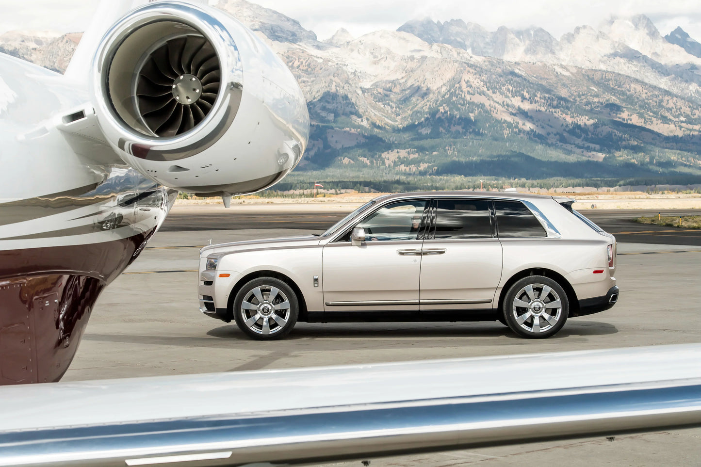 Silver 2019 Rolls-Royce Cullinan SUV parked on airport tarmac near private jet with mountains in background