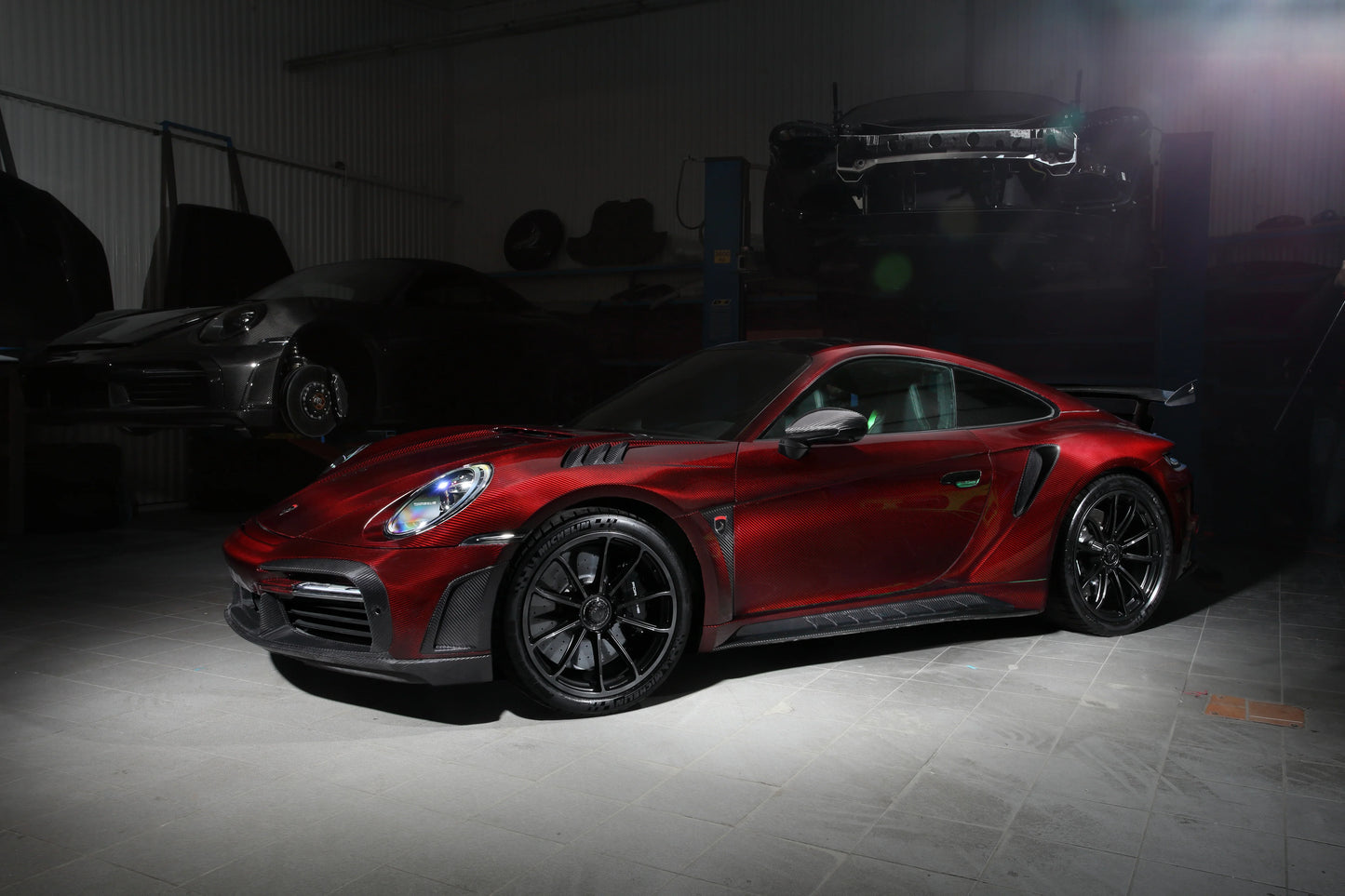 Red Porsche 911 Turbo S with carbon fiber details in a dimly lit workshop with another car lifted in the background