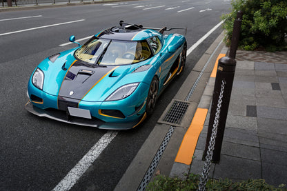 Teal and black Koenigsegg sports car parked on urban street near sidewalk with yellow curb markings